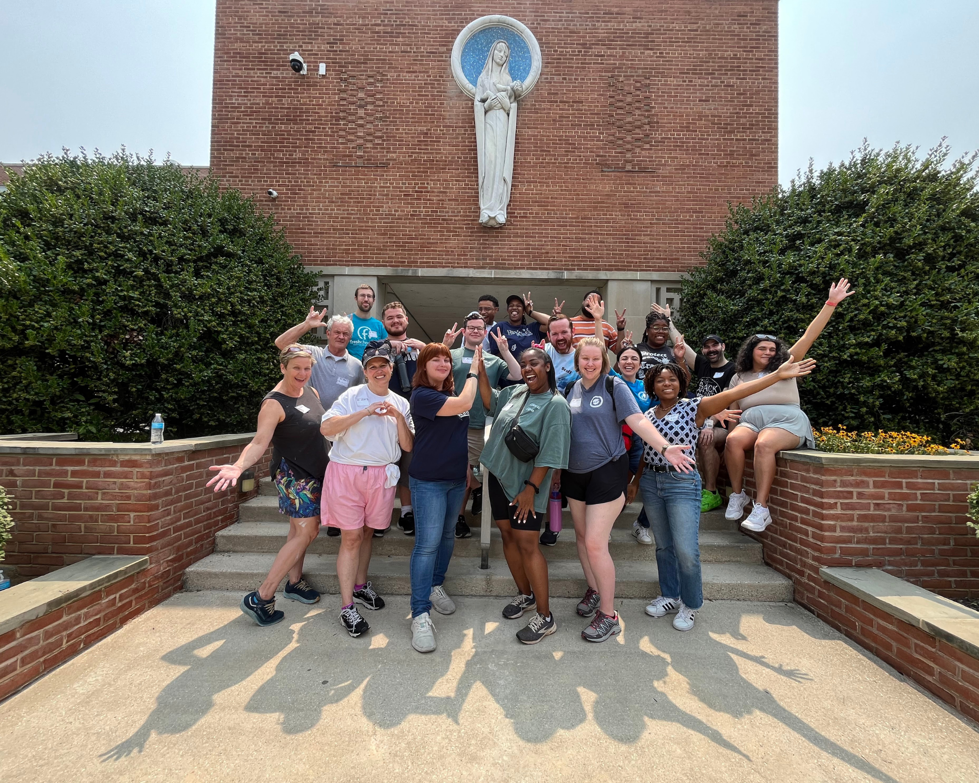 Group of volunteers stand in the sunlight with arms extended, directly outside St. Ann's Center.